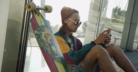 Laughing young man dressed like a hipster in round glasses sits with his skateboard on the floor and watches funny video on his smartphone