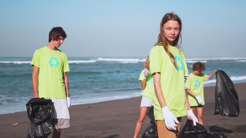 Four volunteers in green t-shirts with image recycle, collect garbage on beach, looking on camera with bags of collected garbage. Volunteering and recycling concept. Environmental awareness copy space Royalty-Free Stock Footage #1016631817