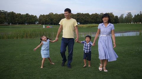 Positive multinational parents with two joyful little siblings walking holding hands along green park lawn while spending weekend together. Happy asian family taking a walk outdoors on summer day.