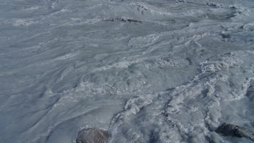 Slow-motion wide pan up from dirty water flowing in the Matanuska River outside of Anchorage, Alaska, to green forest below mountain peak with blue sky and clouds.