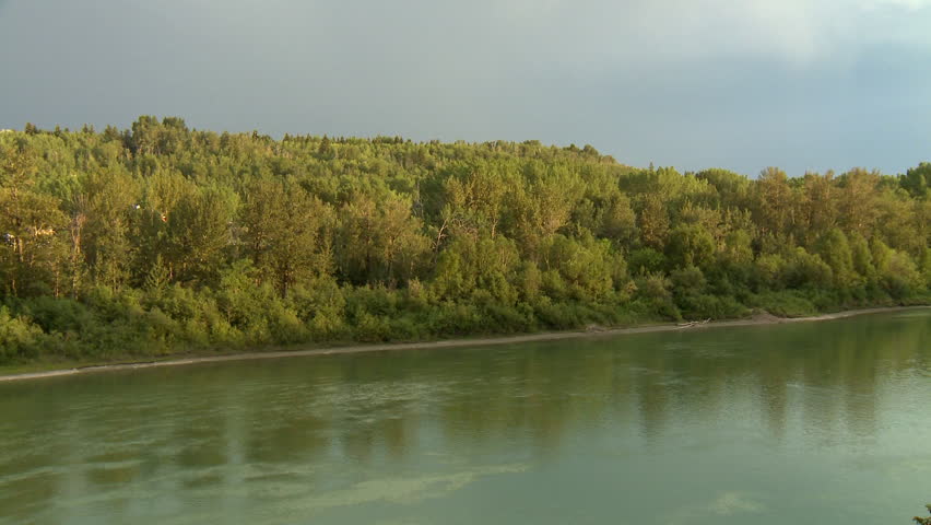 North Saskatchewan river flows past sunlit trees under a darkening sky. Shot in Edmonton, Canada.