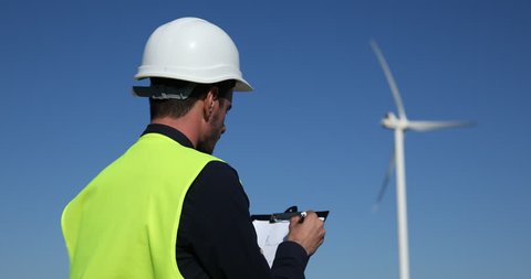 Master Engineer Man Make Notes in Works Documentation Looking Up at Wind Turbine