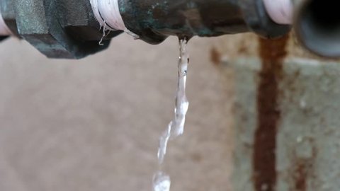 Water dripping from a leaky pipe next to a building. White plumbers tape applied to pipe. Constant stream of water coming from a pipe. Beige, neutral textured wall in background.
