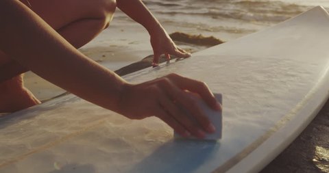 Close up shot of surfer girl waxing surfboard on beach at sunset