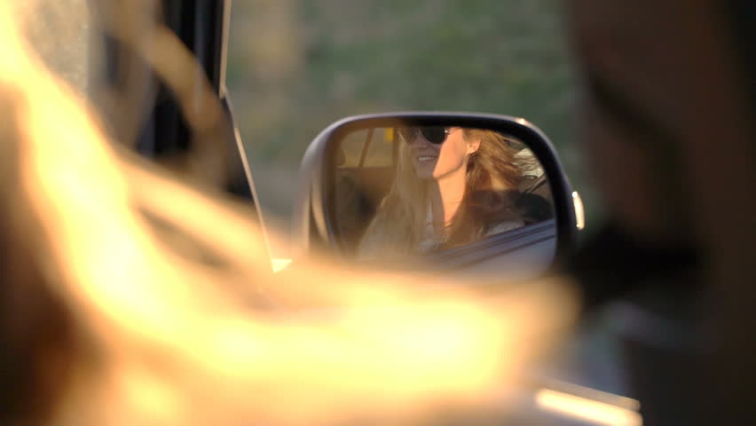 A young woman enjoys traveling in a car with an open window in the rays of the sunset and reflection in the rearview mirror. Slow motion.