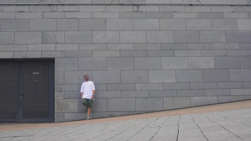 Guy in white T-shirt is standing by the gray wall boy is passing by on the skateboard