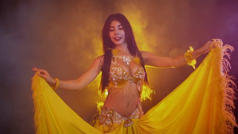 Seductive sexy traditional oriental belly dancer girl dancing on yellow neon smoke background. Woman in exotic costume with feathers sexually moves her semi-nude body.