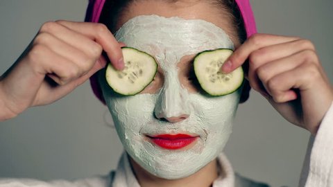 Close up of a girl with a face mask and a towel on her head that closes her eyes with cucumbers on a gray background. Face care concept. Beautiful girl with cosmetic mask on her face