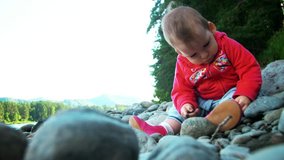 Pretty little girl is sitting on a bank of mountain river and playing with stones. Little baby sit on river bank. Father's day