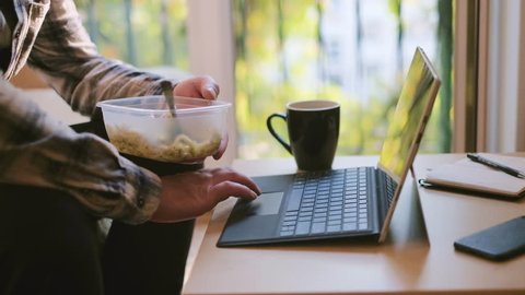 Man workaholic eating pasta from a plastic lunch box while he is working on laptop at home office, no break, static shot of employee sitting on couch, 4K UKD