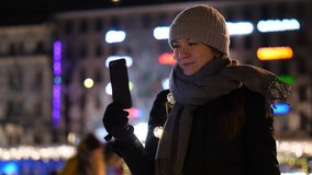 Positive woman have video call from ice rink, hold smartphone and glowing garland in second hand, speak with someone by internet. Tourist lady celebrate Christmas and share emotions and expressions