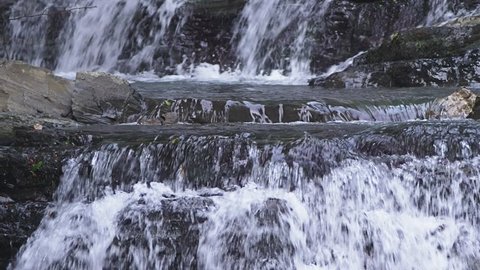 close-up. Waterfall on rocky mountain. Water stream quickly fall down and create white bubbling foam. Stone waterfall in mountain forest. 4k, slow motion