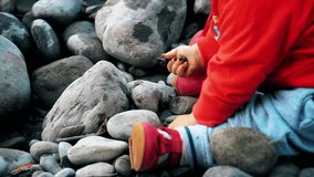 Pretty little girl is sitting on a bank of mountain river and playing with stones. Little baby sit on river bank.
