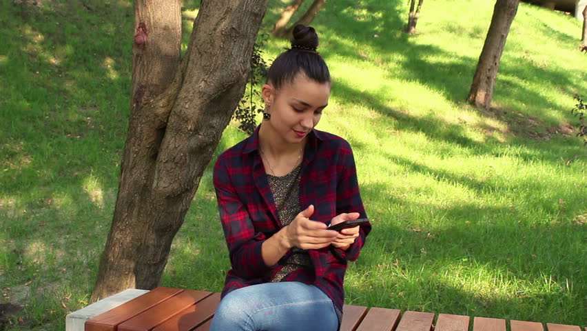Young beautiful girl in a plaid shirt is sitting on a bench in the park, smiling and writing a message on the internet chatting on her smartphone.