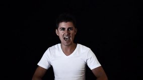 Young good looking sporty male with big blue eyes and stylish haircut dressed in white t-shirt is emotionally singing and showing hand heart shape in front of camera in studio on black background.