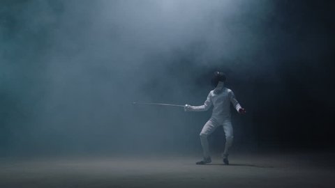 Professional fencer in protective uniform practicing maneuvers with sword alone in dark studio . Fencing demonstration on black smoked background . Shot on Arri Alexa cinema camera in slow motion .