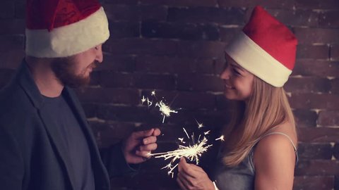 Man with beard and woman with long hair in New Year hats looking at each other, holding Bengal lights, smiling and touching for nose  holding Bengal lights
