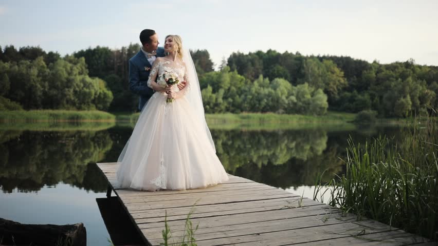 Smiling beautiful young wedding couple stand hugs near lake love bride dress groom outdoors white married woman celebration romantic beauty female man people suit husband kiss slow motion