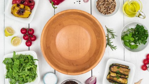 Fresh salad with grilled vegetables in wooden bowl with food ingredients on white wooden rustic table. Top view, 4k stop motion animation.