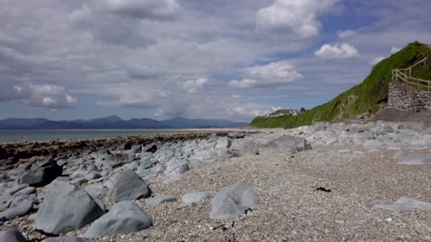 A slow Right to Left smooth Drone track across a rocky beach to darker rocks with distant mountain in the distance in Wales in the UK - 5 second version