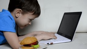 Child blogging in internet, boy with laptop at home on the couch, eating a hamburger, looking at the computer, watch cartoons or talking with friends he eat burgers and look on displayed on the screen