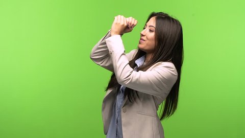 Young business woman on green chroma key background making the gesture of a spyglass, is observing something that is far away