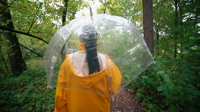 Young pretty woman in yellow raincoat walking in autumn park or forest alone. Girl with transparent umbrella. Rainy weather, nature background.