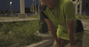 Tired woman in fitness wear resting after training.Side view.Summer evening or night.Industrial green city.Urban runner cardio healthy activity workout.4k slow motion 60p video