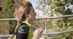 Young blond woman in fitness wear running training on aerial walkway.Side following view.Summer sunny day.Industrial green city.Urban runner cardio healthy activity workout.4k slow motion 60p video