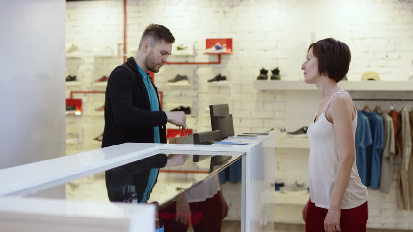 Cheerful customer is using a credit card to pay for purchased clothes while standing at cashier's desk. Shop assistant is accepting transaction and giving paper bags. Royalty-Free Stock Footage #1016822746