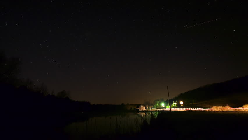 Time Lapse of the Starry Night Sky by a Pond with Cars Driving By