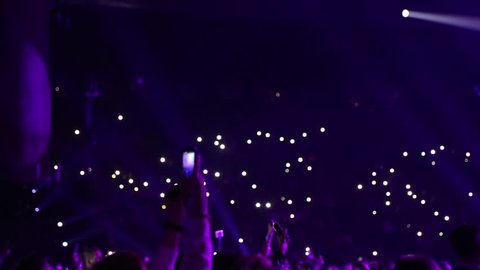 Defocused spectators in hall with lights sitting in dark. Audience is shining with flashlights of phones. Concert hall with spectators.