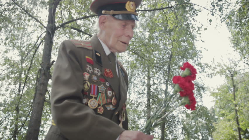 An elderly gray-haired veteran of the great Patriotic war and world war II in uniform with many badges and orders puts red flowers to the monument. Steadicam shot