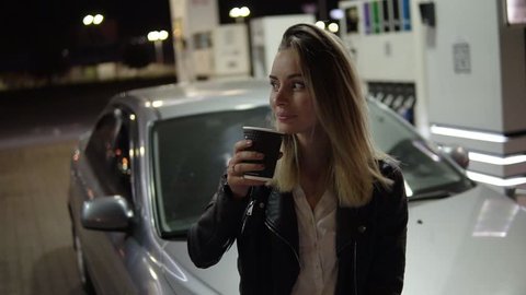 Having rest. Pretty smiling woman leaning on the car and holding black paper cup, drinking coffee. Front view
