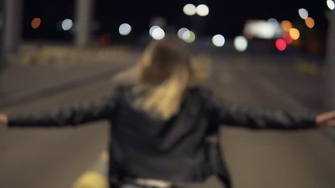 Happy blonde woman having fun outdoors at the nighttime. near the airport building. Running, immitating plane, jumping on bumpers. Wearing jeans and black jacket. Rare view
