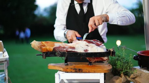 Chef who cuts a slice of ham or dried meat, of excellent quality, with the knife according to tradition. Concept of ham, italian food. Catering outdoors. 4K UHD