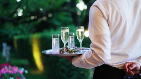 Waiter and champagne and water glasses on a tray in restaurant or event party meets clients and guests at the entrance. Catering staff. 4K UHD.