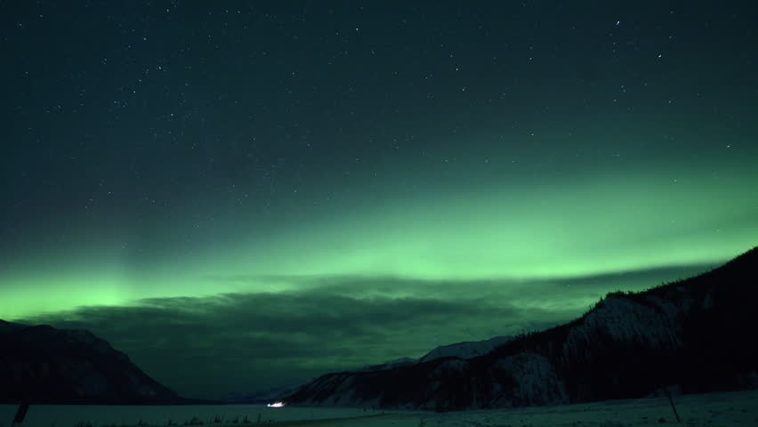 Time lapse shot of aurora borealis over lake and mountains against star field at Northern Rocky Mountains Provincial Park