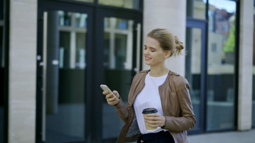 Cheerful young businesswoman surfing her mobile phone while walking down street and stopping to text back in messenger Royalty-Free Stock Footage #1016890579