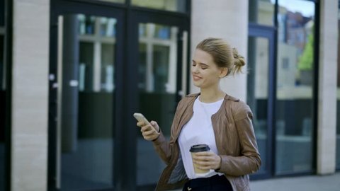 Cheerful young businesswoman surfing her mobile phone while walking down street and stopping to text back in messenger