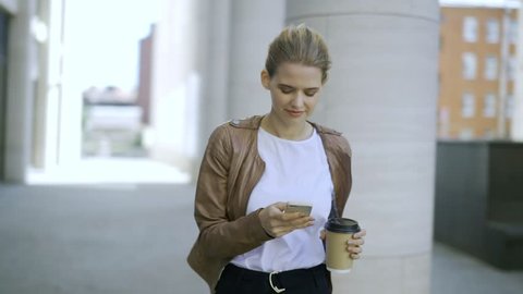 Panning shot of young smiling woman with paper coffee cup standing in street and texting on phone or using mobile application