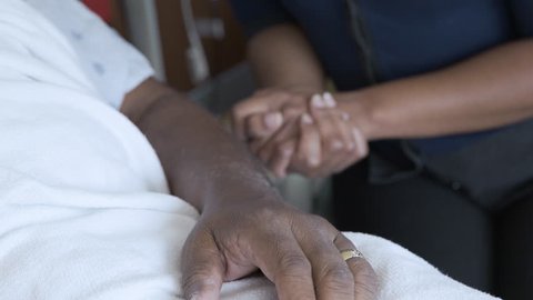 Handheld shot of daughter touching father's hand lying on bed at hospital ward