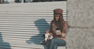 Pretty caucasian woman with dog jack russell terrier sitting on bench and drinking coffee. Cute little puppy 4K video shooting