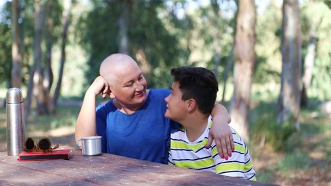 Portrait of a happy sick woman fight with cancer with family's help. Mother and son sitting at the park bench