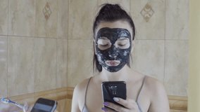 The girl cleans her teeth and sits on the Internet from the phone. She has a black mask on her face. The camera makes a reflection in the mirror. Real-time video.
