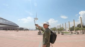 A young man, tourist standing with a backpack and in a hat and shooting a video by smartphone near the Communication Tower of Santiago Calatrava and the Olympic Ring of Barcelona, Spain