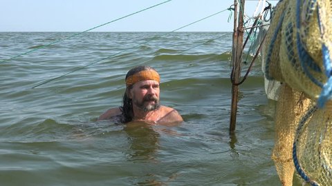 Mature male fisherman in kerchief with beard dives into water to repair fishing nets in ocean. Fishing nets are dried in open sea near shore with a small storm