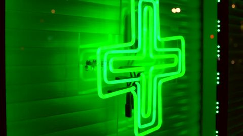 This panning night time video shows a bright green neon medical weed cross on an urban marijuana dispensary's store front window.