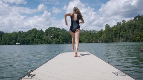 Girl runs and jumps off a floating dock on a lake in slow motion. Wide angle of the millennial woman jumping in the air and splashing into the water embodies freedom and healthy lifestyles. 