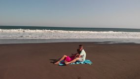 4K aerial flying video of young couple on the beach with black volcanic sand at sunset time. Bali island.